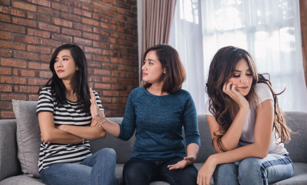 Three women sitting on a sofa, with one gesturing between the others.