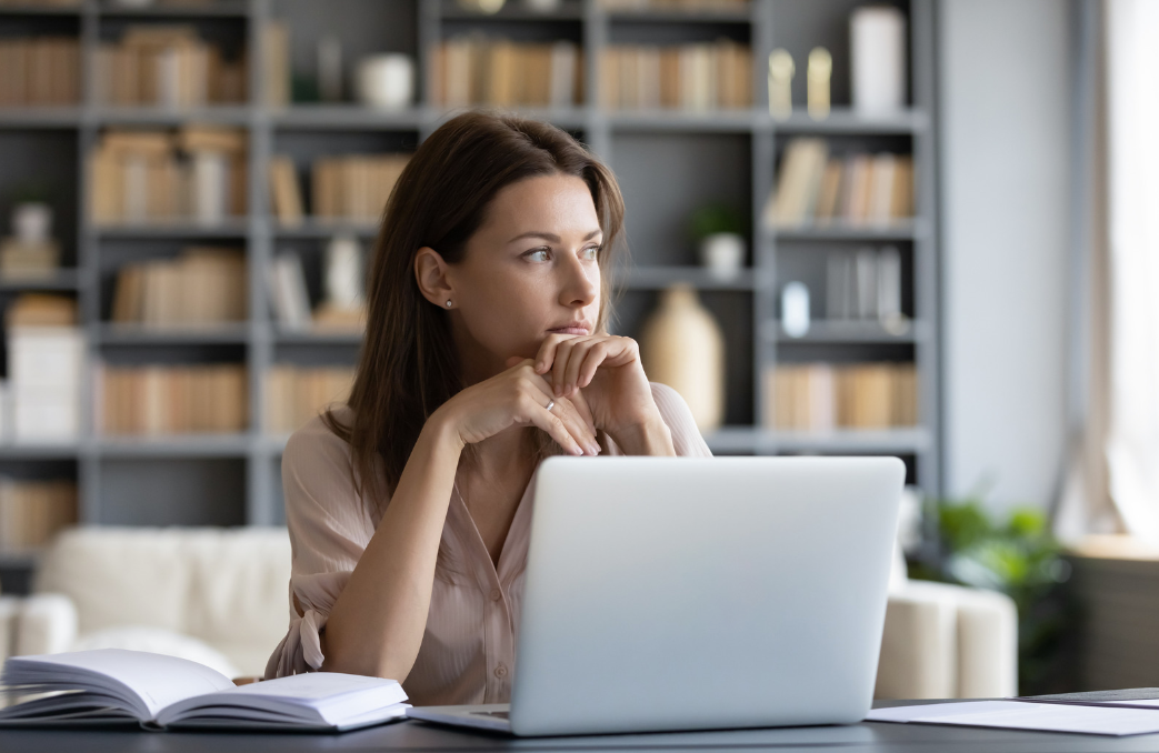 Woman seated at a table with an open laptop and book in a bright room.